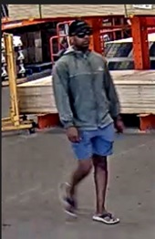 Photo de surveillance du suspect portant une casquette de baseball noire, des lunettes de soleil, un manteau gris à manches longues muni d’une fermeture à glissière et un short bleu.