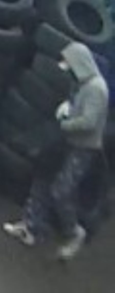 Le suspect debout : Un suspect portant des chaussures blanches, un pantalon bleuté et un chandail gris dont le capuchon recouvre un chapeau à rebord tient un sac noir et marche devant un tas de pneus.  